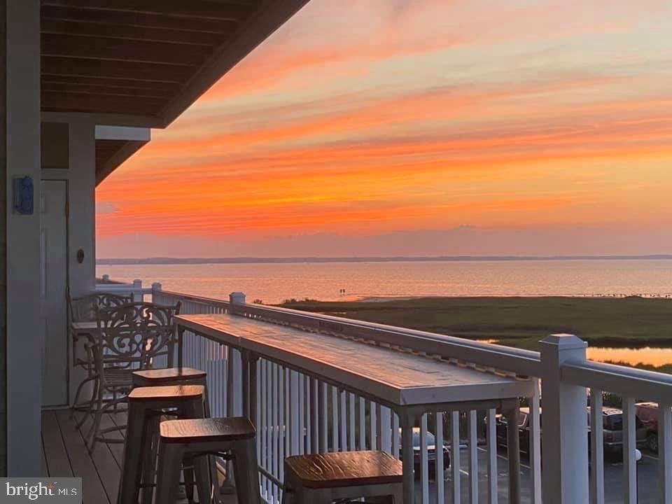 A balcony offers a serene view of a vibrant sunset, with orange and pink hues reflecting on the water. Bar stools are positioned along the railing, inviting relaxation. The horizon features a peaceful landscape, with gentle waves and grassy shores visible.