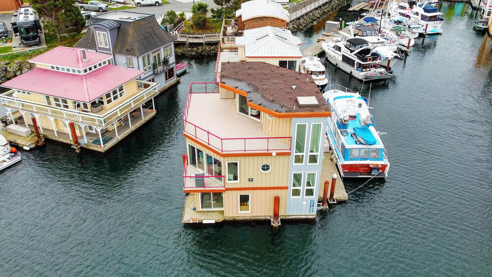 A modern float home is shown from an aerial perspective, surrounded by water and neighboring boats. The multi-level structure features large windows and a prominent deck, offering views of the marina and other nearby homes. The vibrant colors enhance its contemporary design.