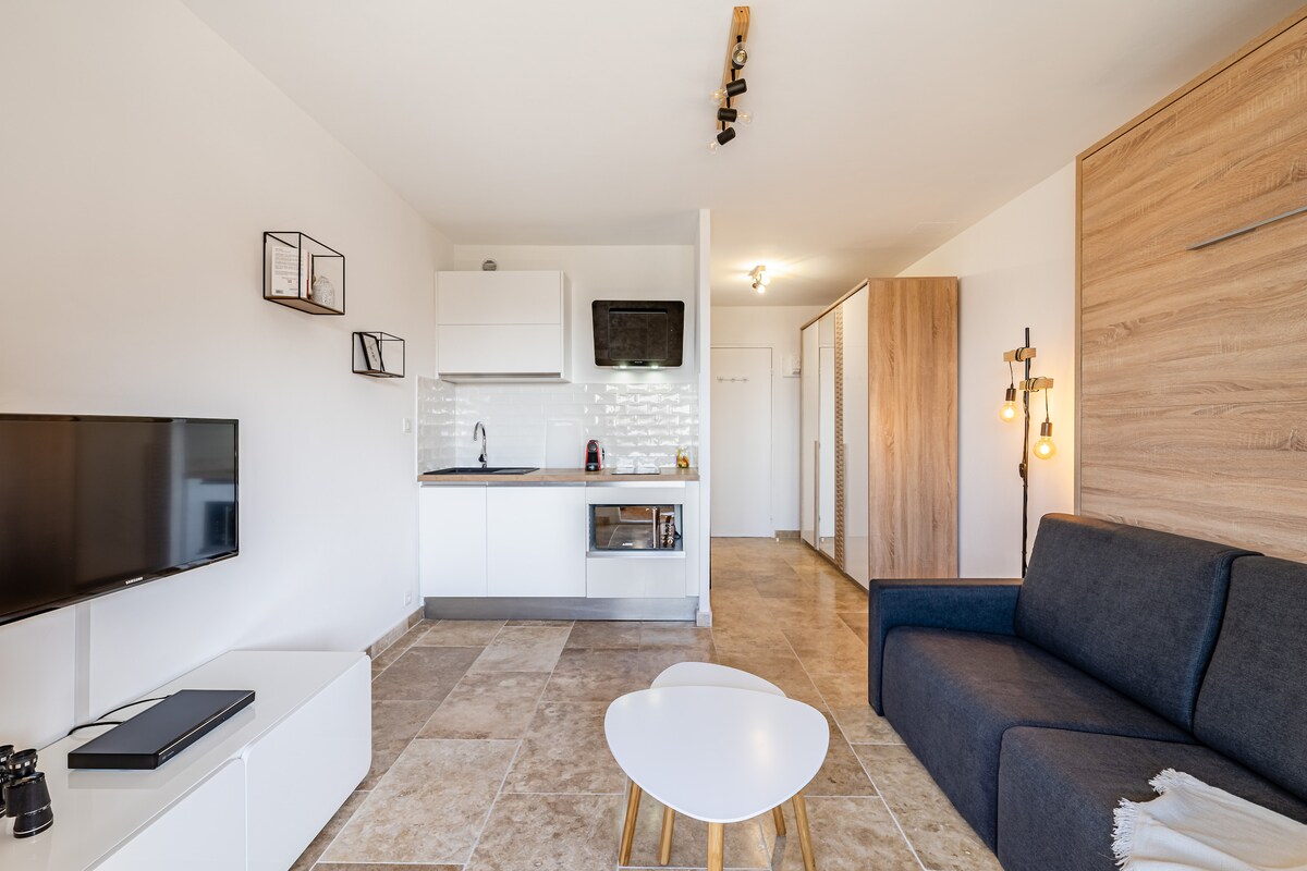 A modern studio features a compact kitchen area with white cabinets and sleek appliances. A plush sofa faces a flat-screen television, and a small, circular table is situated on the tiled floor. Warm lighting accents the wood paneling, enhancing the clean design.