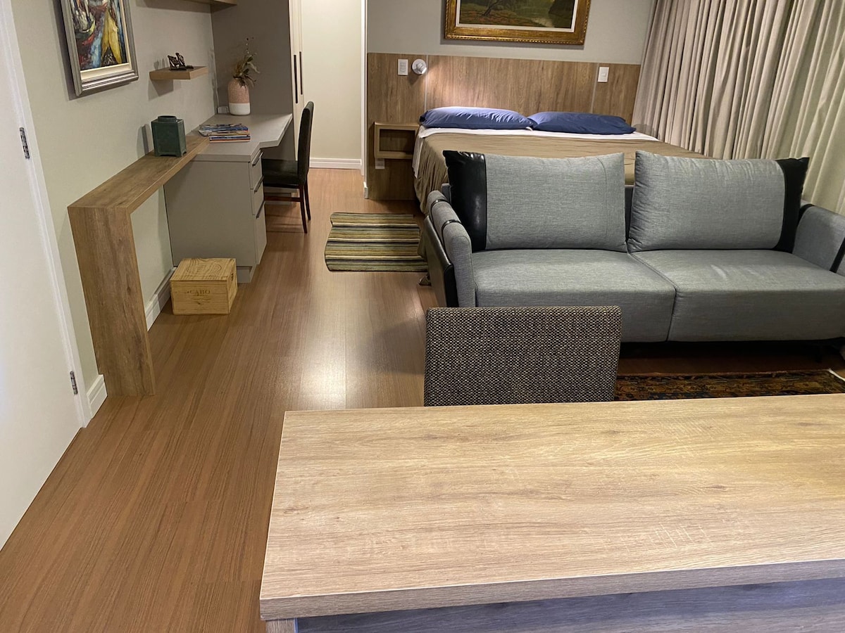 A spacious living area features a modern gray sofa and a wooden coffee table. A low dresser and desk provide additional functionality, while a bed is positioned in the background. Neutral tones and wood accents create a cohesive and inviting ambiance.