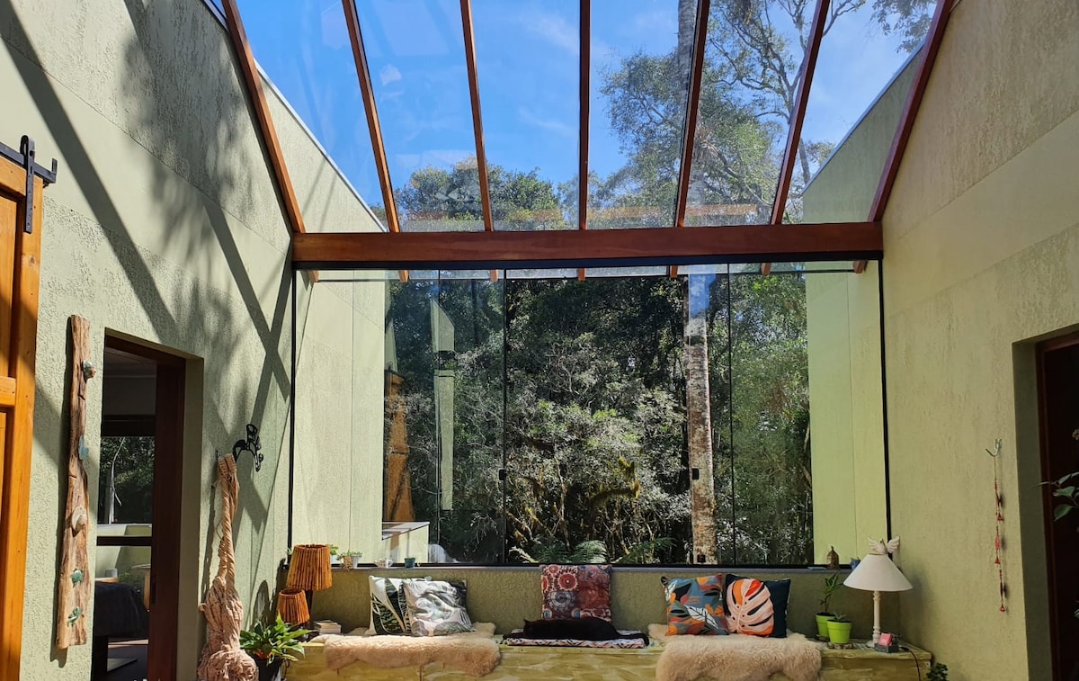 A spacious area is highlighted by a glass-paneled roof, allowing natural light to fill the room. Soft seating features plush textiles and is arranged near expansive windows showcasing views of the surrounding greenery. Decor elements include decorative cushions and a lamp.