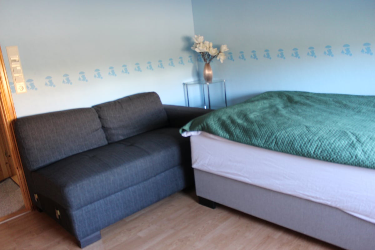 A sleeping area features a spacious bed dressed in a green blanket and a comfortable sofa in a soft blue room. A clear vase with white flowers sits on a small table, adding a touch of decor to the area.