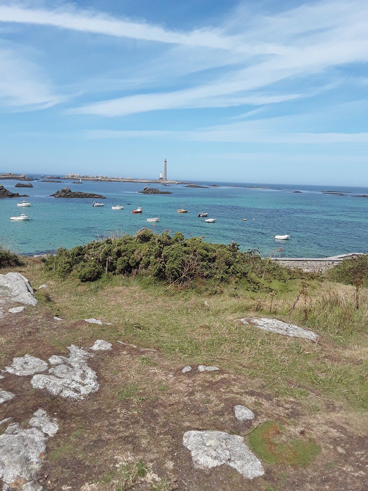 A coastal scene features a clear blue sky above a serene body of water dotted with small boats. In the distance, a lighthouse stands on a rocky outcrop, framed by rugged shoreline and patches of greenery. The landscape conveys a sense of tranquility.
