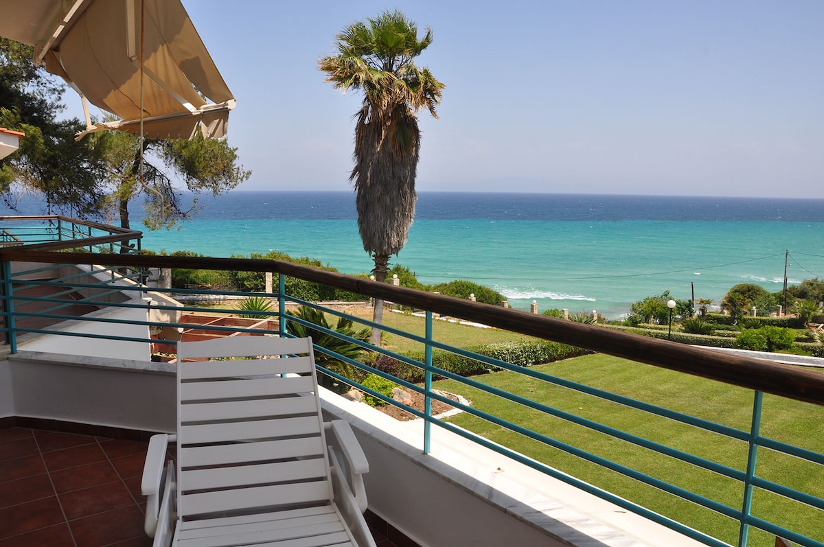 A balcony view features a sun lounger overlooking a vibrant sea and sky. A tall palm tree adds greenery to the scene, while manicured lawns and coastal plants frame the picturesque location, inviting relaxation and enjoyment of the natural surroundings.