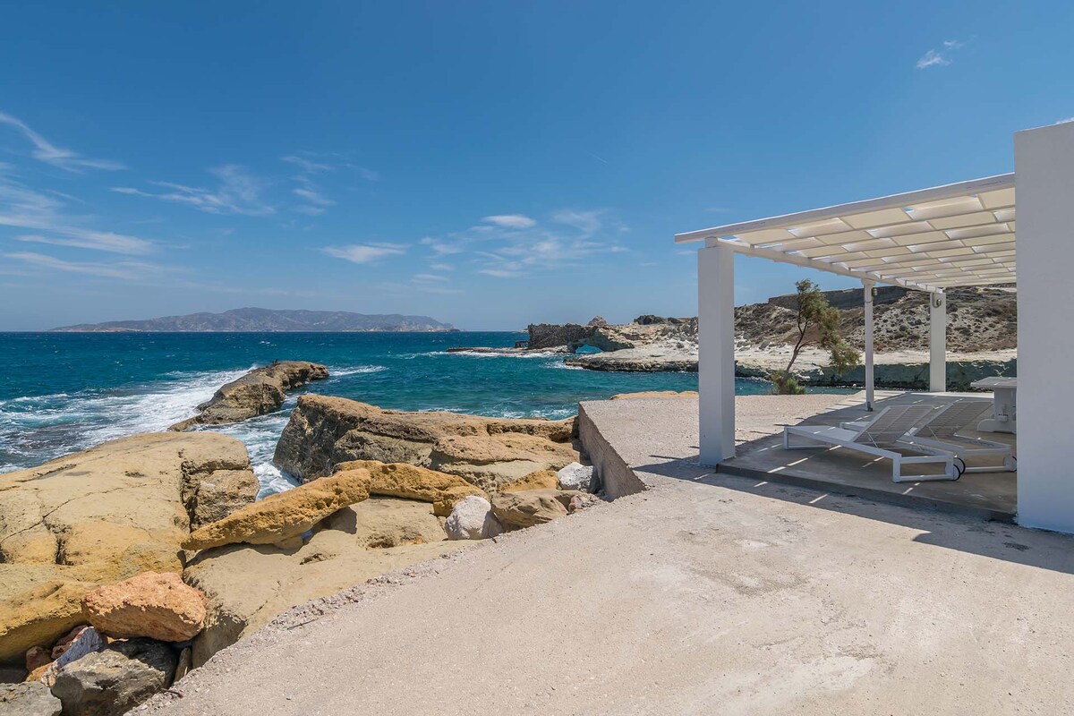 An outdoor terrace is shown, offering views of the rugged coastline and the sea. Clear skies are present above, and the gentle waves of the water can be seen lapping against the rocky shore. A shaded area with ample lounging space is visible.