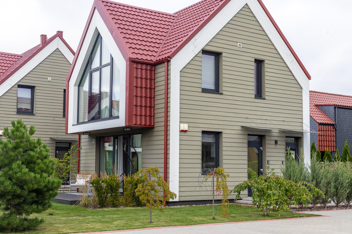The modern apartment building features a combination of light wooden panels and bold red roofing. Large, angular windows invite natural light, while surrounding greenery adds a touch of nature. A welcoming terrace is visible at the entrance, providing an outdoor relaxation area.