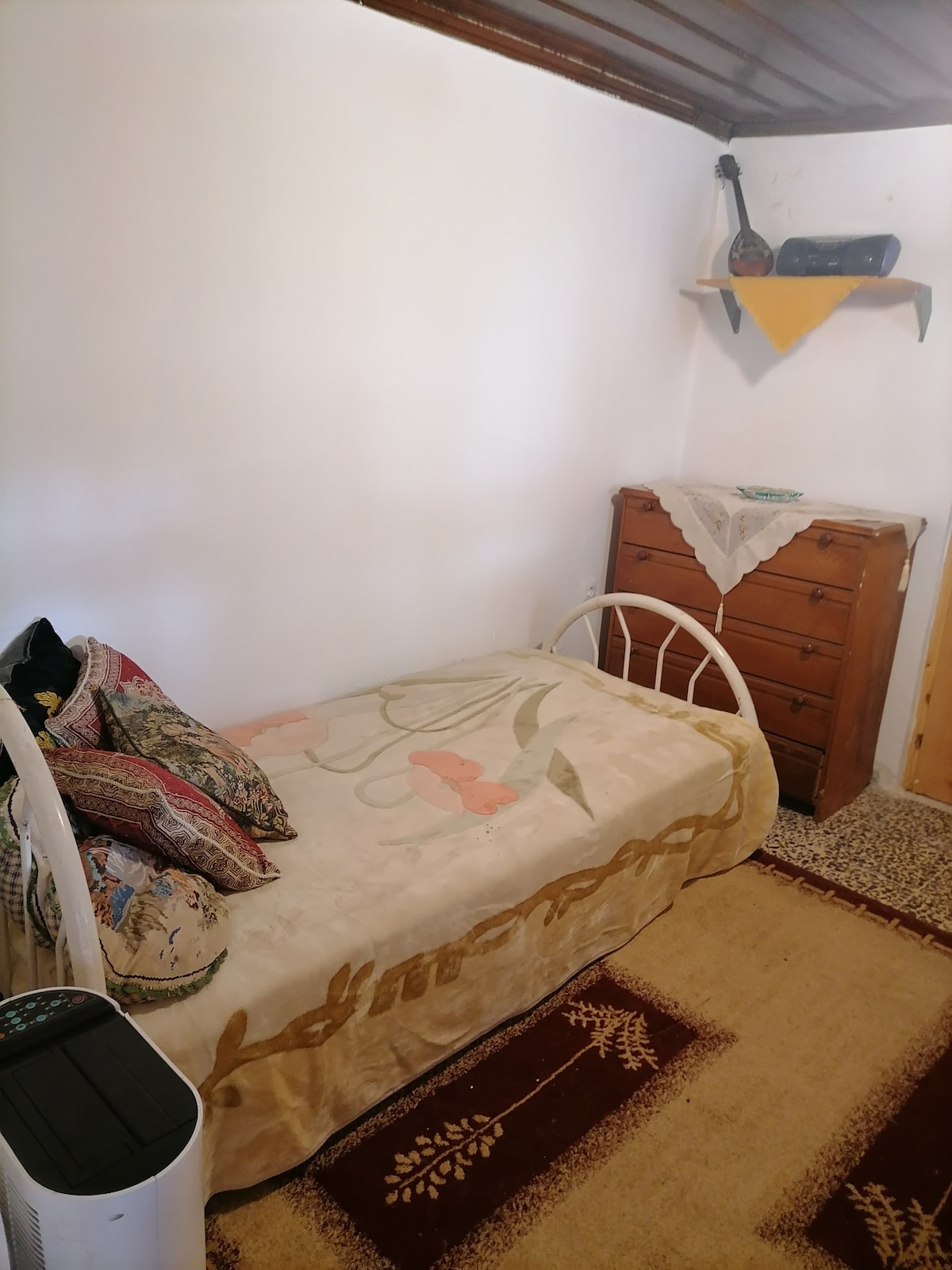 A simple bedroom features a single bed adorned with a floral patterned blanket and several decorative pillows. A wooden dresser with multiple drawers stands opposite the bed, topped with a lace cloth and a vintage object. A patterned area rug covers the floor.