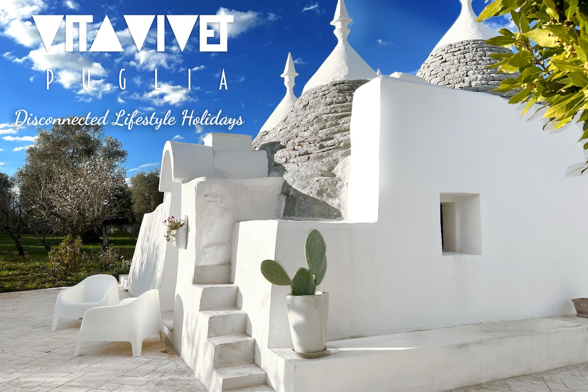 A traditional trullo showcases a white-washed exterior, complete with a distinctive conical roof. Steps lead to an entrance, while minimalist seating is arranged outdoors. The surrounding area features lush greenery, creating a serene atmosphere ideal for relaxation.