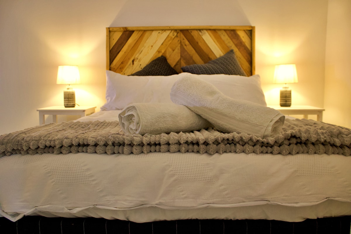 A spacious bed is presented with neatly rolled towels placed on top. Soft lighting from two bedside lamps creates a gentle glow, highlighting the textured throw blanket draped across the bed. The headboard features a rustic wood design, adding warmth to the room.