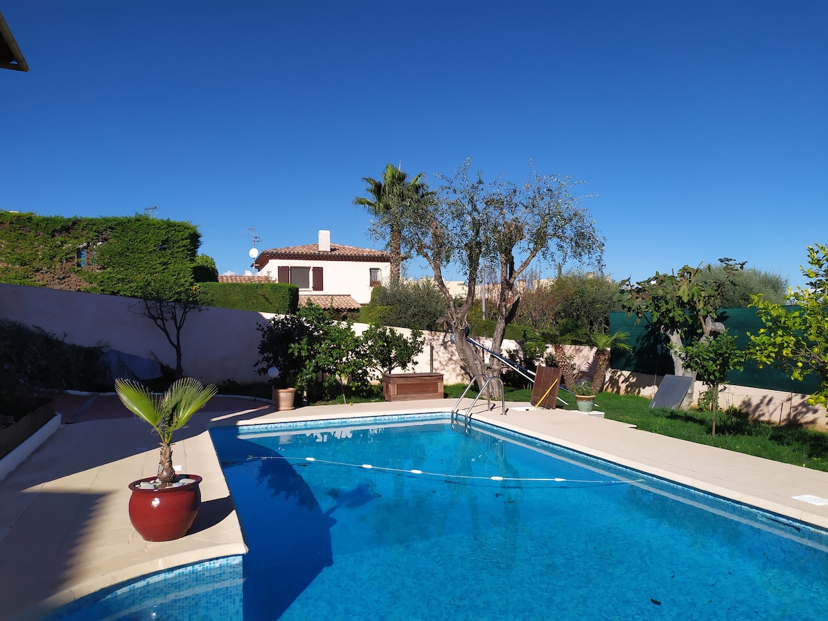 A clear blue pool reflects the bright sky, surrounded by well-maintained gardens featuring tropical plants and trees. Lounge chairs are arranged on the pool deck, offering a space for relaxation. The villa's exterior can be seen in the background, enhancing the tranquil ambiance.