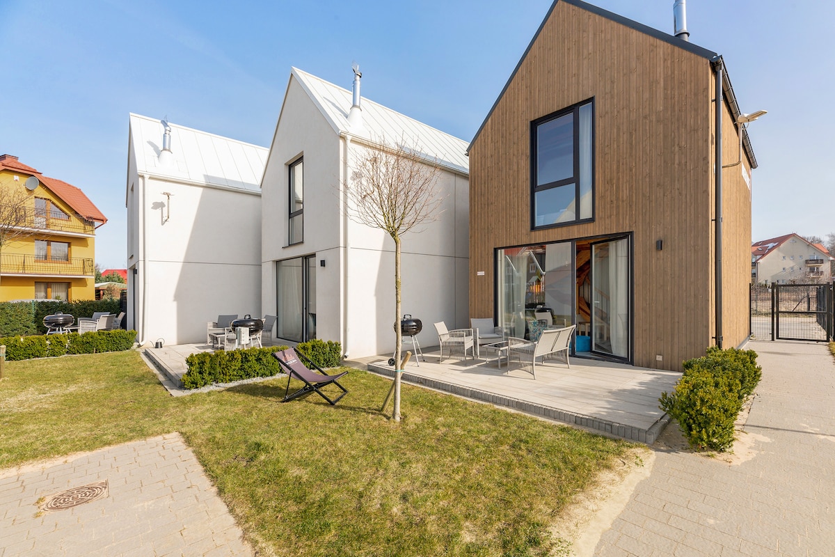 Two modern houses are positioned side by side, featuring a mix of white and wooden facades. A wooden terrace is set with outdoor furniture and a grill. A small tree and green shrubs line the edge of a lawn, enhancing the outdoor space.