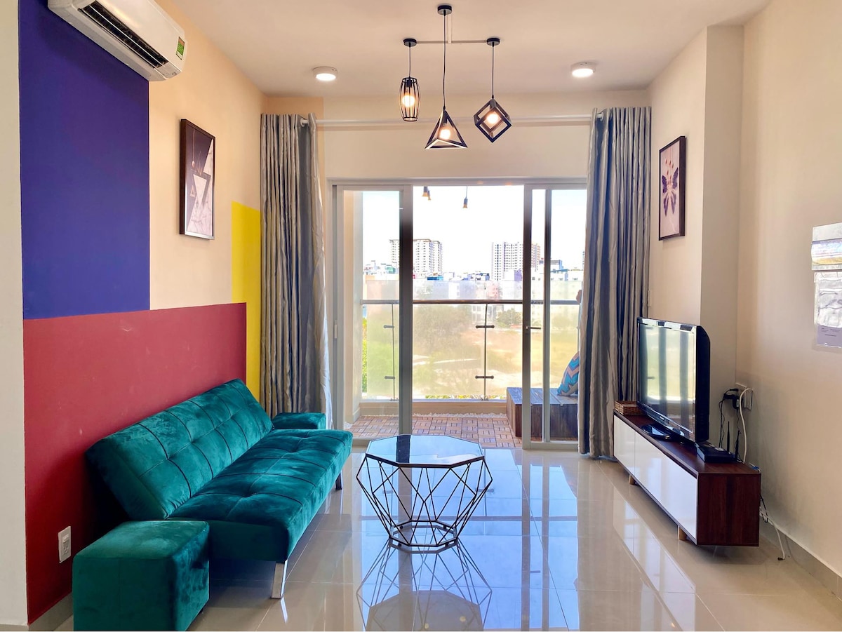 A modern living area features a teal sofa, a geometric coffee table, and a television on a console. Natural light fills the space through large glass doors that lead to a balcony. Color blocks in red and yellow on the wall add visual interest.