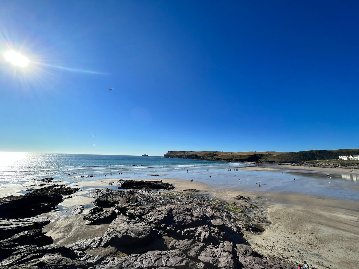 A wide view of a sandy beach under a clear blue sky, with gentle waves lapping at the shore. The rocky outcrops are scattered along the foreground, while a few figures can be seen enjoying the beach. A distant coastline outlines the horizon.