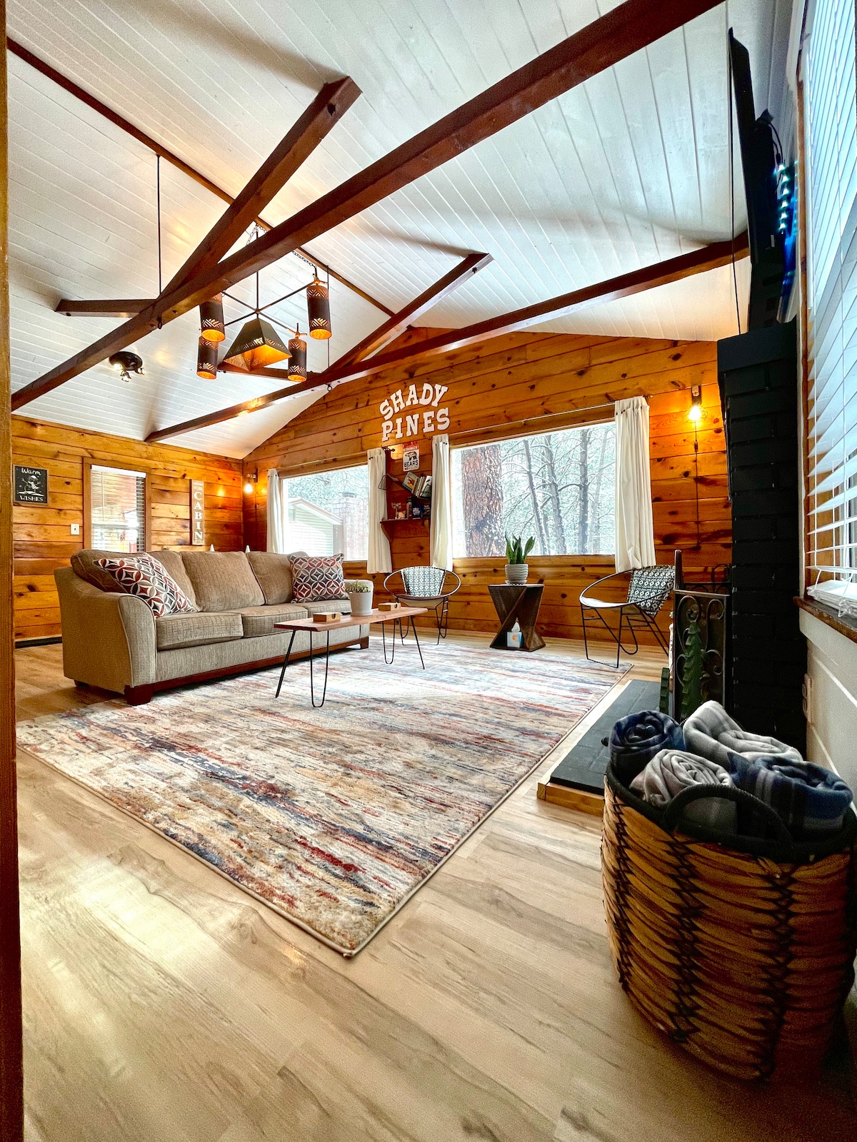 The open-concept living area features wooden walls and a light wooden floor. A comfortable sofa is positioned alongside two chairs and a small table. Natural light enters through multiple windows, illuminating a colorful area rug, with a decorative sign reading 'Shady Pines' on the wall.