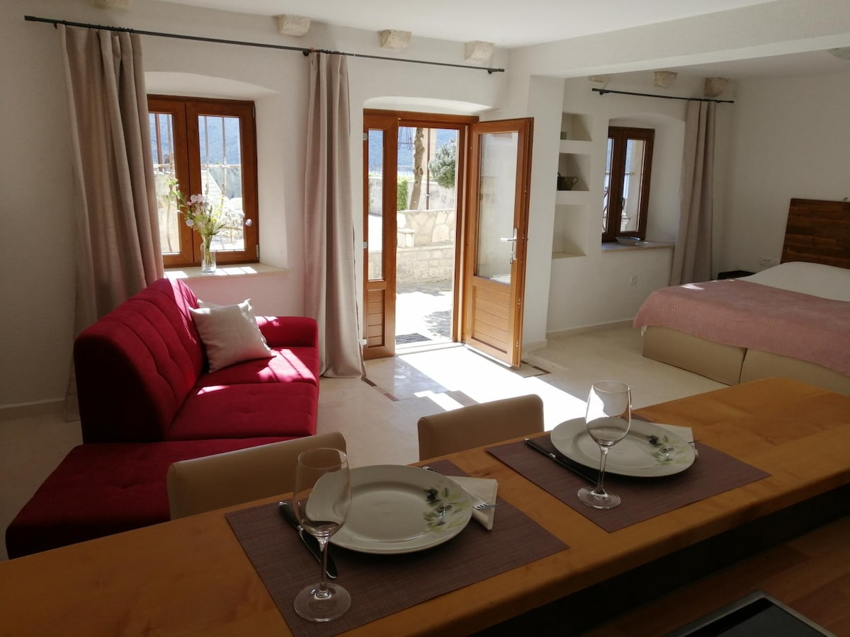 An inviting living space includes a plush red sofa and a spacious bed, with large windows allowing natural light to fill the area. A dining space is set with two plates and glasses, while pale curtains soften the atmosphere.