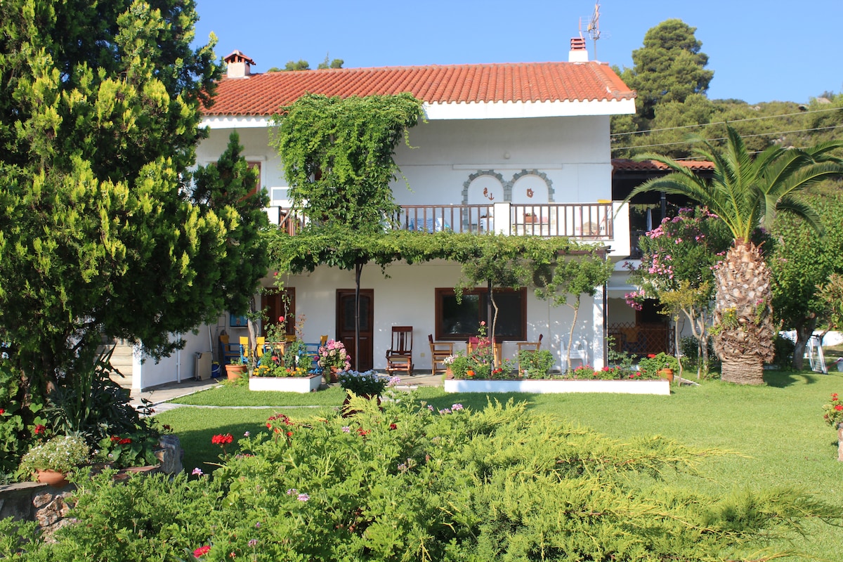 A two-story building is surrounded by lush greenery and vibrant flowers, offering a touch of nature. A balcony extends from the upper level, enhancing the outdoor aesthetic. The well-maintained garden features a variety of plants, contributing to a serene and inviting setting.