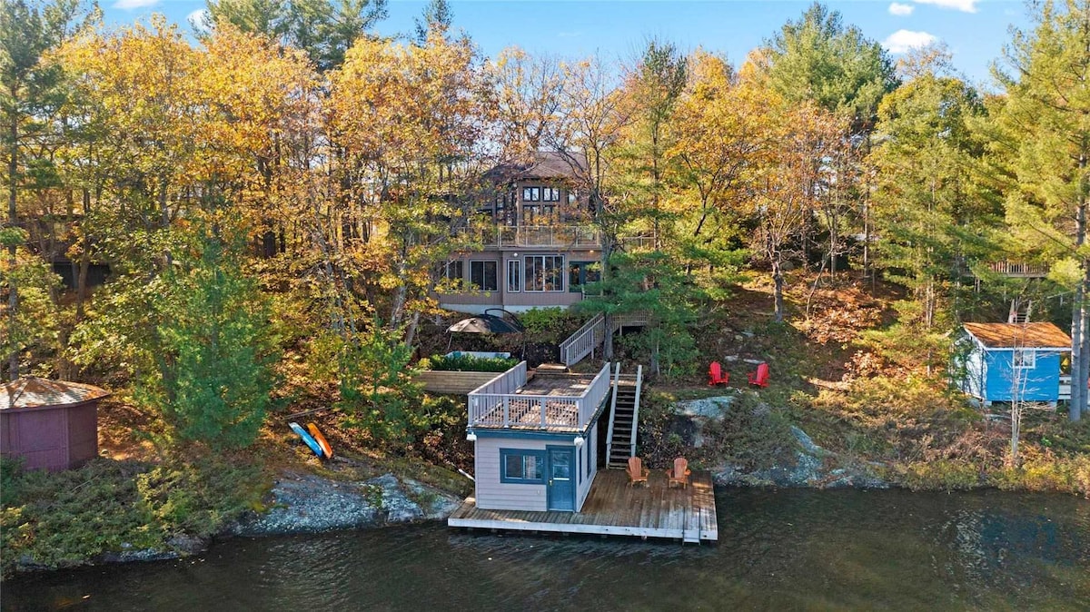 The cottage is nestled among mature trees in a vibrant autumn landscape, with colorful foliage visible. A spacious deck extends to the waterfront, featuring two Muskoka chairs and a staircase leading to the dock. Kayaks are positioned nearby, ready for use.
