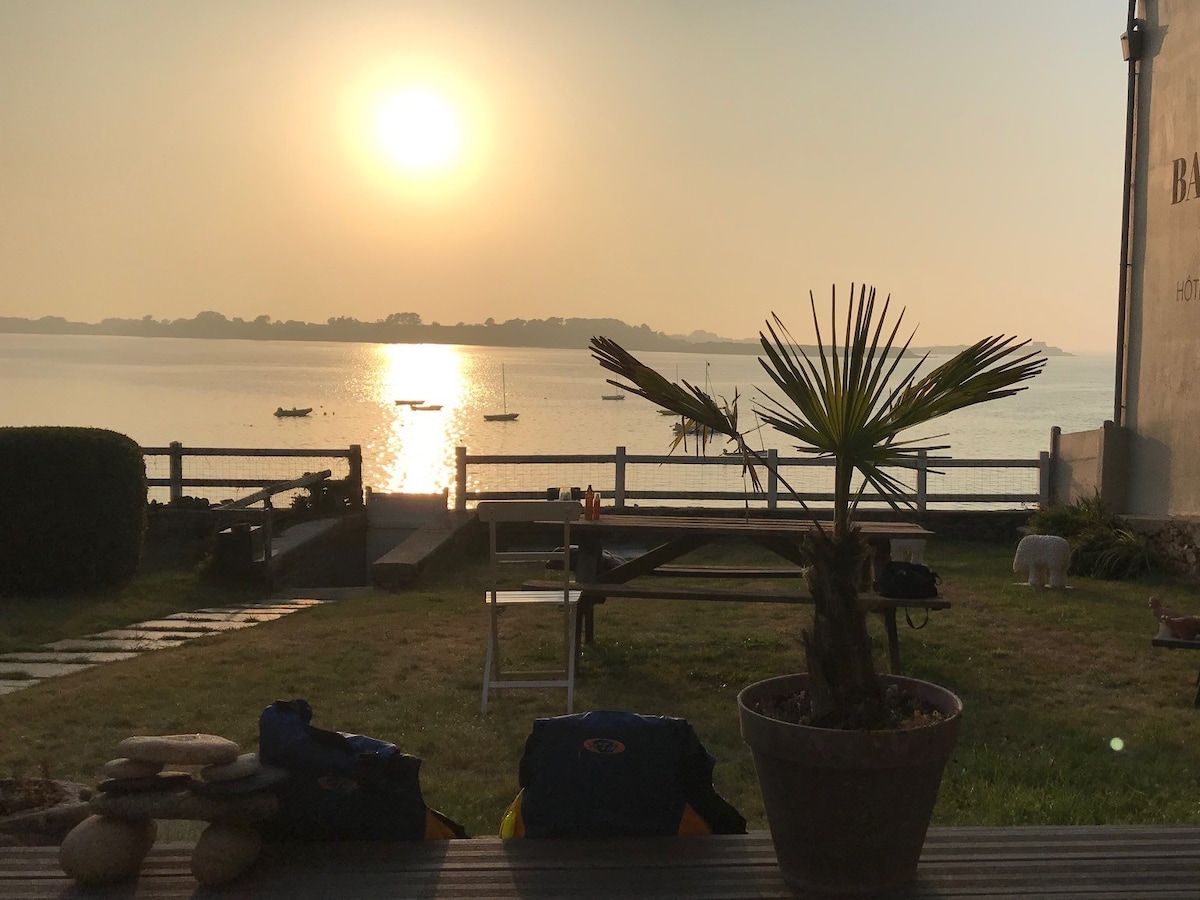 A serene outdoor view features a calm body of water reflecting the golden hues of the setting sun. In the foreground, a potted plant is placed on a wooden table, with stones arranged nearby. Small boats can be seen in the distance, enhancing the tranquil atmosphere.