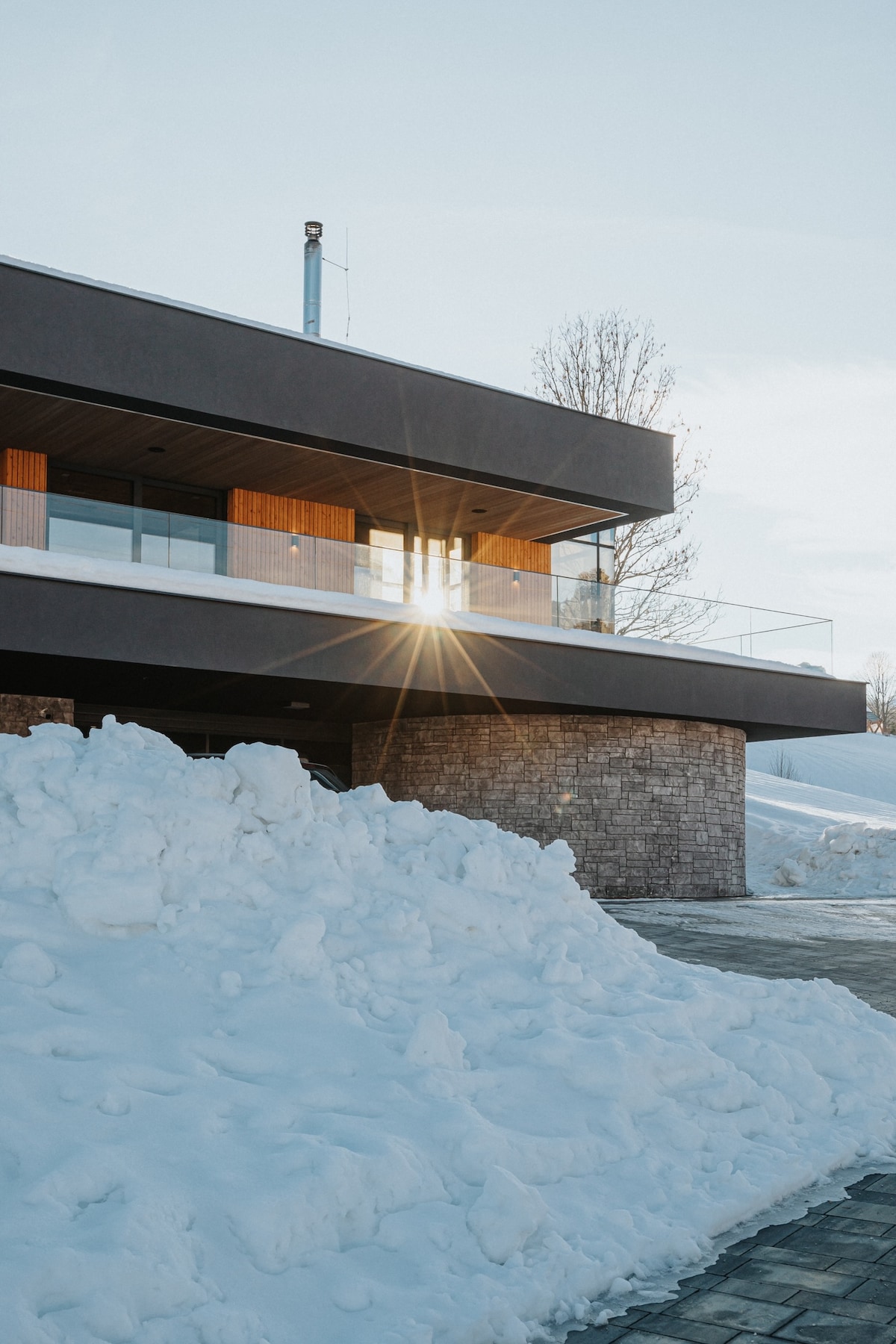 A modern villa is set against a winter landscape, with large windows reflecting sunlight. A mound of snow fills the foreground, while a stone pathway leads to the entrance. The design showcases clean lines and a contemporary aesthetic.