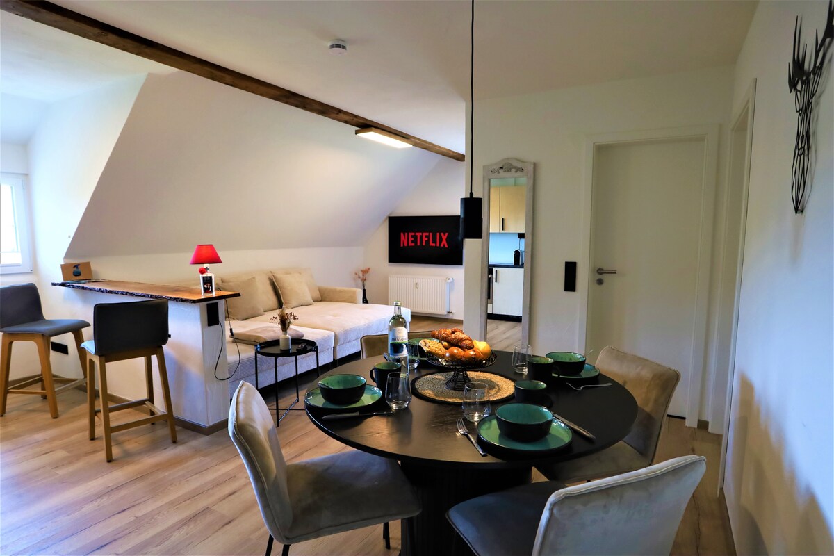 The open living area features a modern dining table set for four, complemented by plush chairs. A comfortable seating area with light-colored couches is nearby, and a wall-mounted television displays the Netflix logo. Natural light enters through a window, enhancing the bright space.