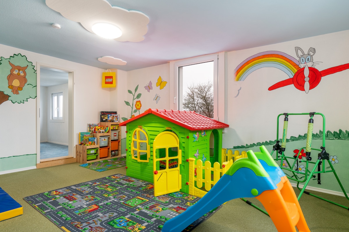 A vibrant indoor play area is filled with colorful toys and murals. A small green playhouse with a red roof is surrounded by a fence, complemented by a slide and climbing structure. The walls are decorated with cheerful illustrations, enhancing the playful atmosphere.