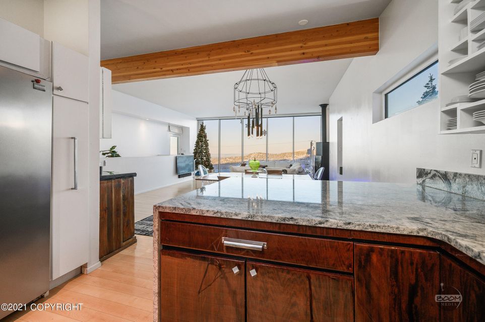 A modern kitchen space features a large island with dark wood cabinetry and a sleek countertop. Large windows provide views of the surrounding landscape. A dining area with a contemporary chandelier is present, while an inviting seating area is visible in the background.