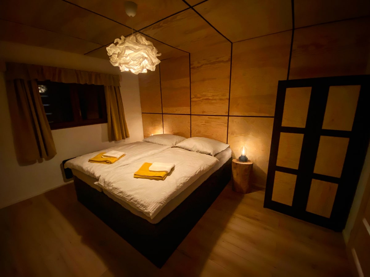 A cozy bedroom features a spacious bed with light bedding, complemented by yellow towels neatly arranged. Soft lighting from a unique lamp casts a warm glow on wooden-panelled walls, while curtains frame a window, enhancing the tranquil atmosphere.