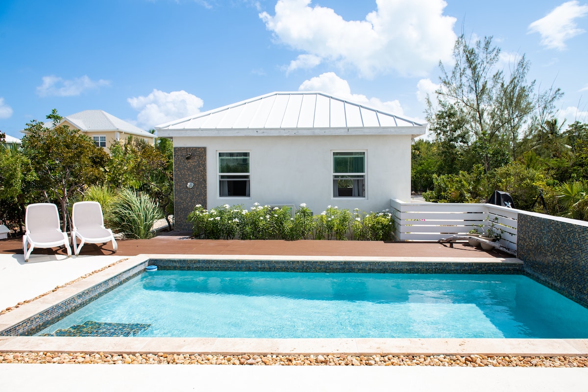 A serene outdoor area features a clear blue pool surrounded by a wooden deck. Two white lounge chairs are positioned beside the pool, complemented by green landscaping and flowering plants. A light-colored house with a metal roof is visible in the background.