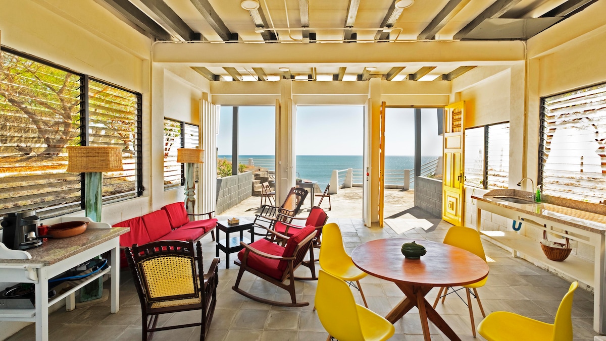 The living area features bright seating options, including red and yellow chairs, alongside a round dining table. Large windows provide ocean views and natural light, while wooden beams and slatted blinds add a modern twist to the design.