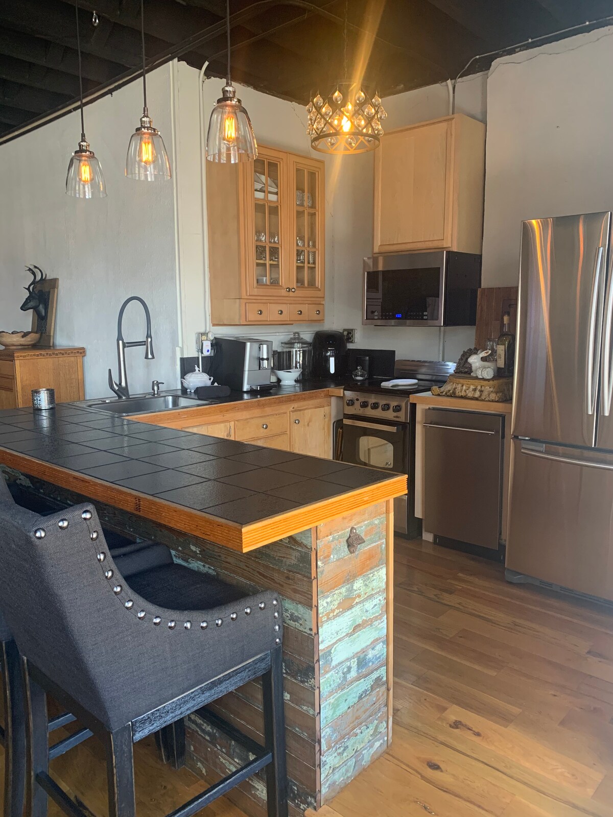 A well-equipped kitchen features wooden cabinetry and a tiled countertop. Stainless steel appliances include a refrigerator and an oven. Three hanging pendant lights illuminate the space. A breakfast bar with dark upholstered chairs offers a place for casual dining.