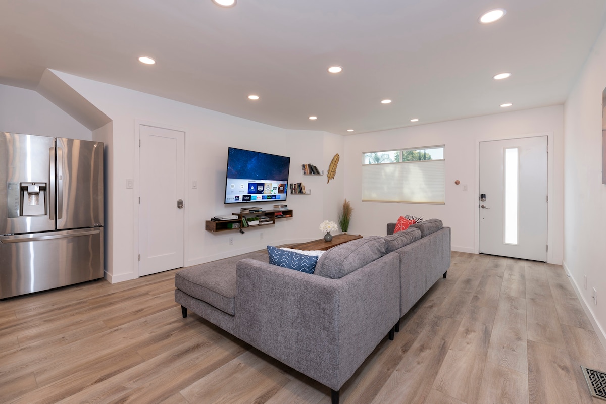 A spacious living room is presented with a large grey sectional sofa facing a mounted television. Light cabinetry and a stainless steel refrigerator occupy one side. Natural light filters through a small window, while a bright front door adds to the inviting atmosphere.