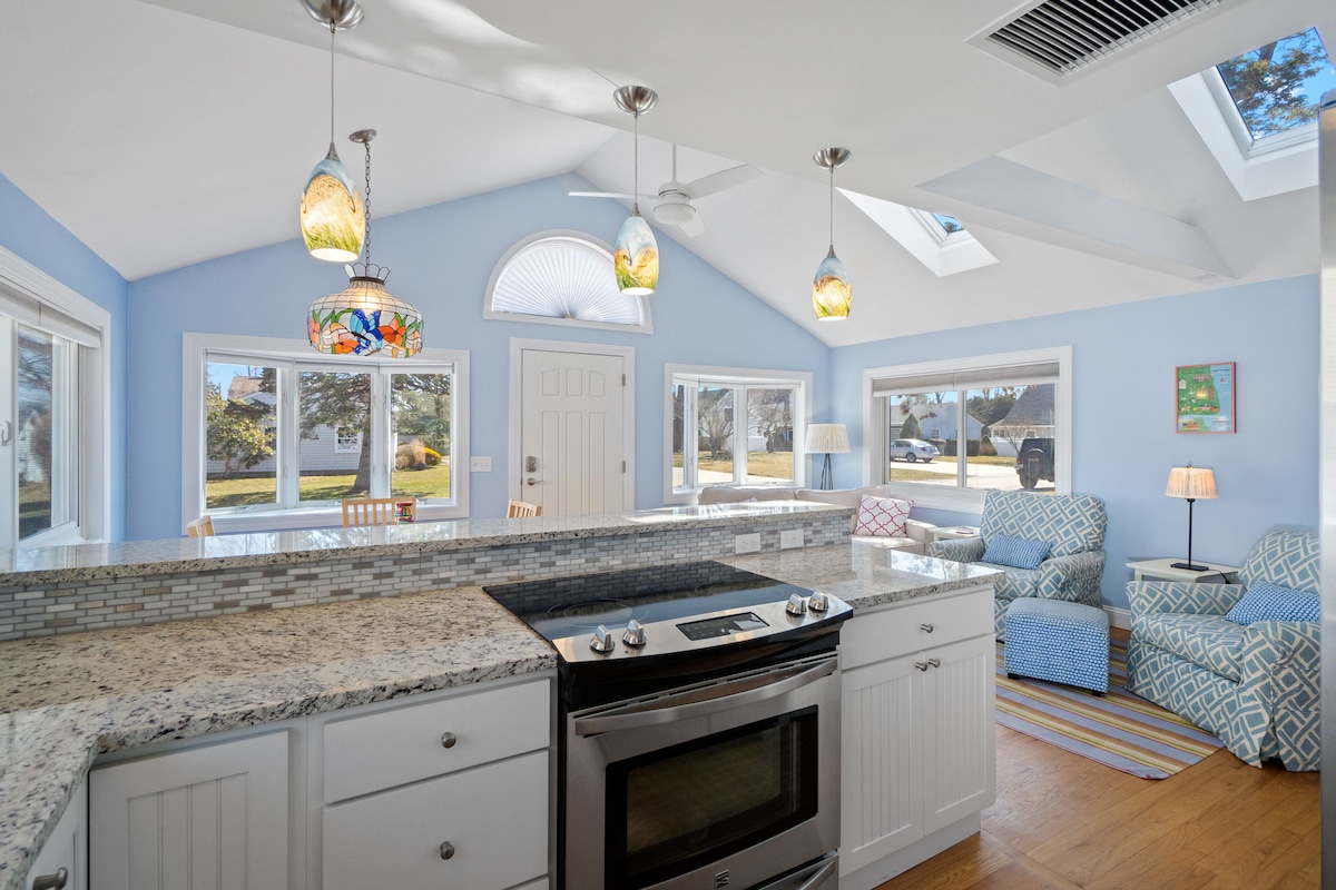 A bright kitchen area is highlighted by granite countertops and a stainless steel oven. Colorful pendant lights hang above, with large windows allowing natural light to fill the space. Comfortable seating with patterned upholstery is visible in the background, offering a welcoming atmosphere.