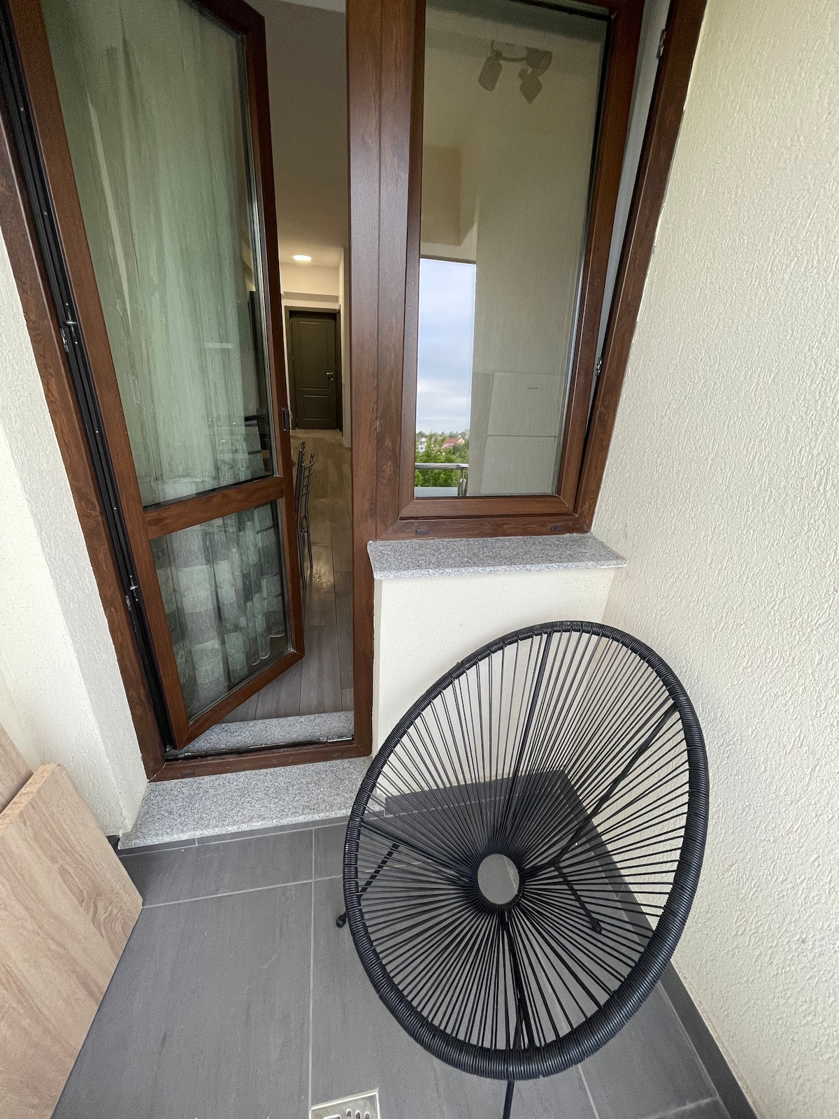 An open balcony is shown featuring a round, black wire chair positioned next to a partially opened glass door. A smooth grey floor is visible, complemented by light-colored walls. The door leads into a well-lit interior space, enhancing the seamless connection between indoor and outdoor areas.