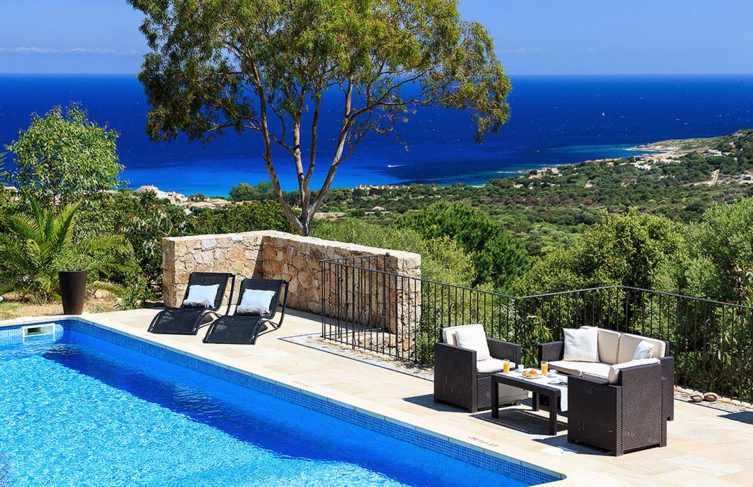 An inviting outdoor pool area features a spacious pool with sun loungers and a cozy seating arrangement. A stone wall adds structure, while the panoramic view of the blue Mediterranean Sea and lush greenery enhance the tranquil setting.
