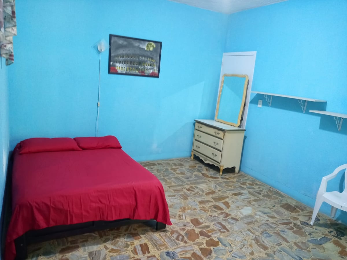 A bright room features a red bedspread over a spacious bed, complemented by a vintage dresser and a wall-mounted shelf. The light blue walls contrast with earthy stone flooring, creating a serene environment. Natural light is introduced by a window with patterned curtains.