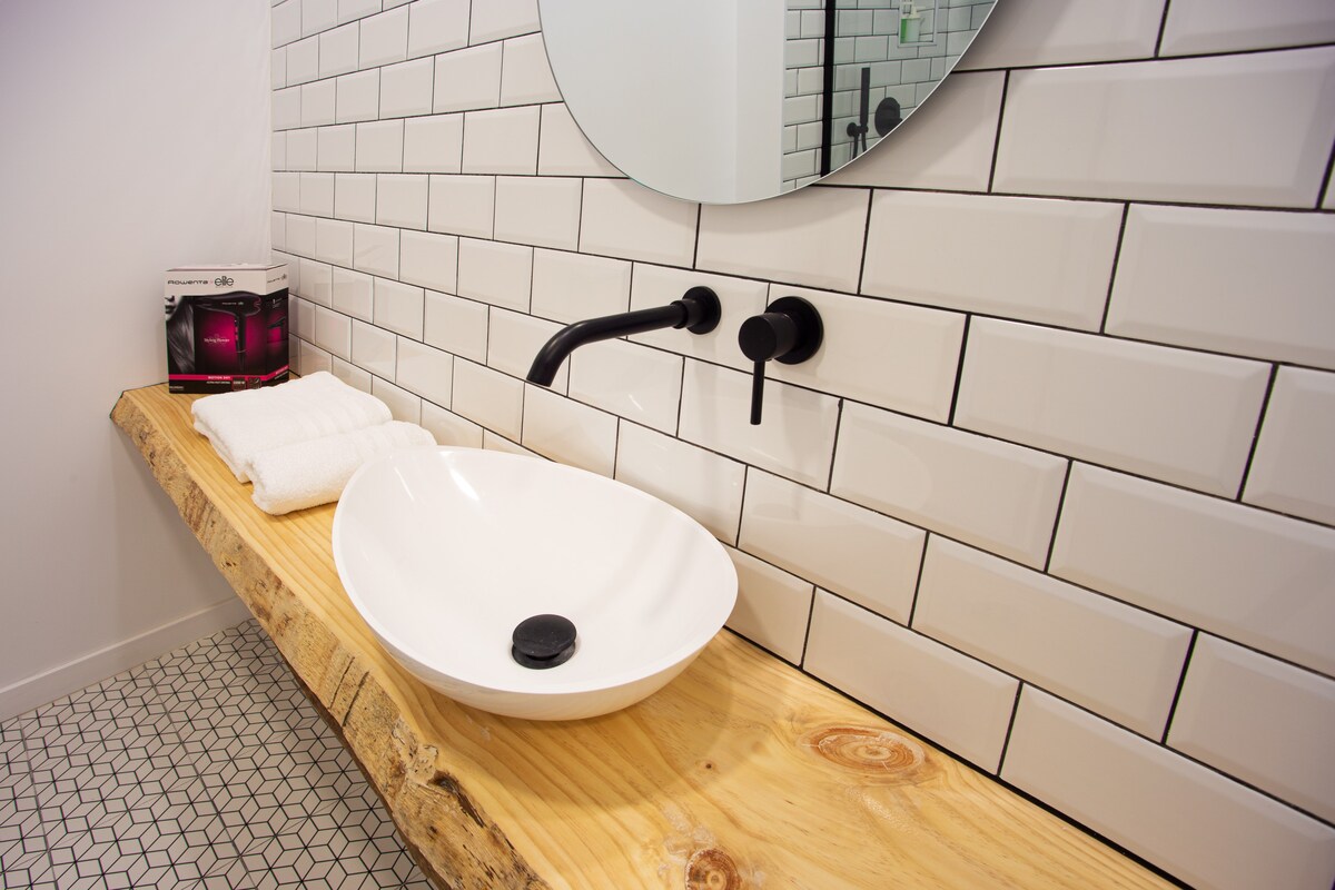 The modern bathroom features a wooden countertop, supporting a sleek white basin. A round mirror is positioned above the sink, while black faucets and fixtures provide a stylish contrast. Soft white towels are neatly arranged nearby, enhancing the clean and minimalist design.