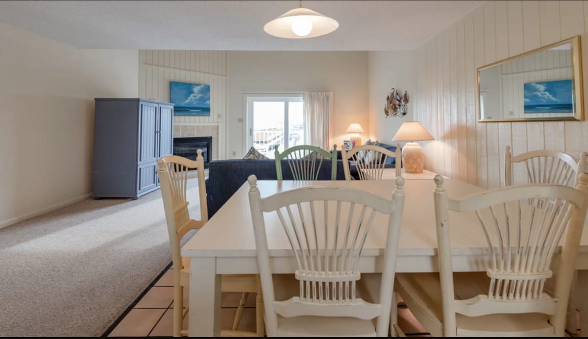 A dining area includes a large white table surrounded by six matching chairs. A comfortable sofa is visible in the background, and a flat-screen TV is placed within a media unit. Natural light is filtered through the balcony doors, enhancing the spacious layout.