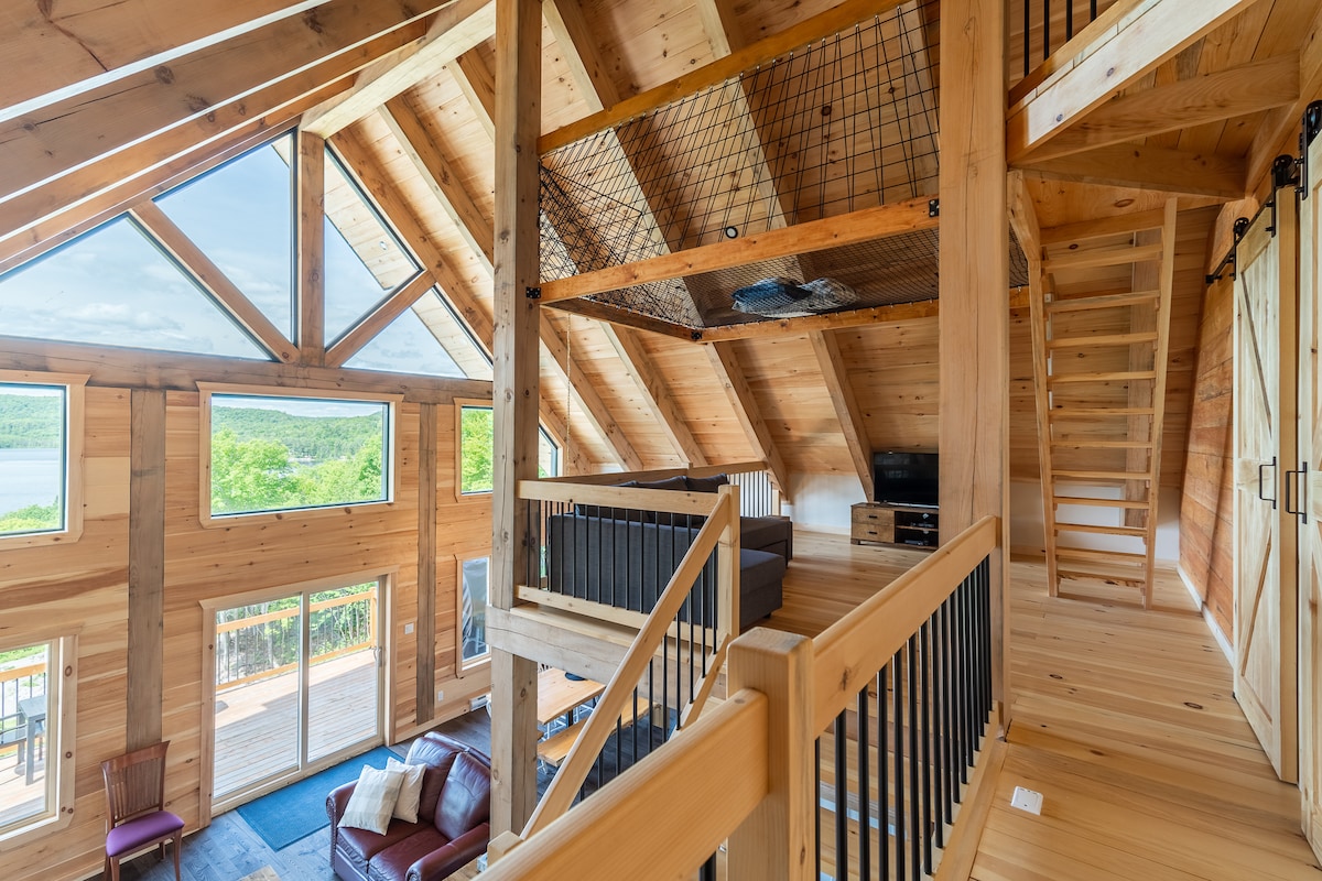 The interior space showcases a spacious design featuring wooden beams and large windows that allow natural light. A cozy seating area with a leather couch is visible, as well as a mezzanine with a hammock overlooking the common area. Stairs lead to an upper level.