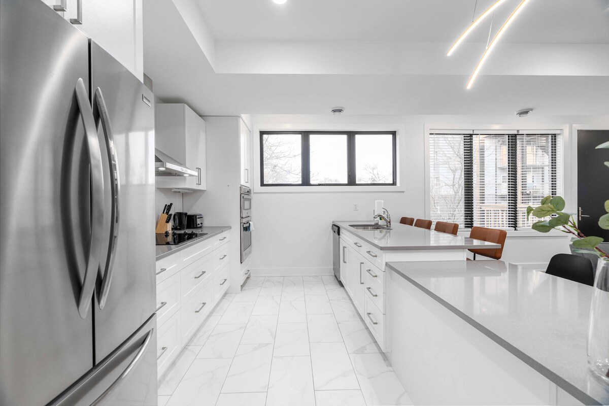 A modern kitchen features sleek white cabinetry and gray countertops. Stainless steel appliances, including a refrigerator and oven, reflect light. A window offers natural light, while three leather chairs are positioned at a nearby countertop, enhancing the social atmosphere of the space.