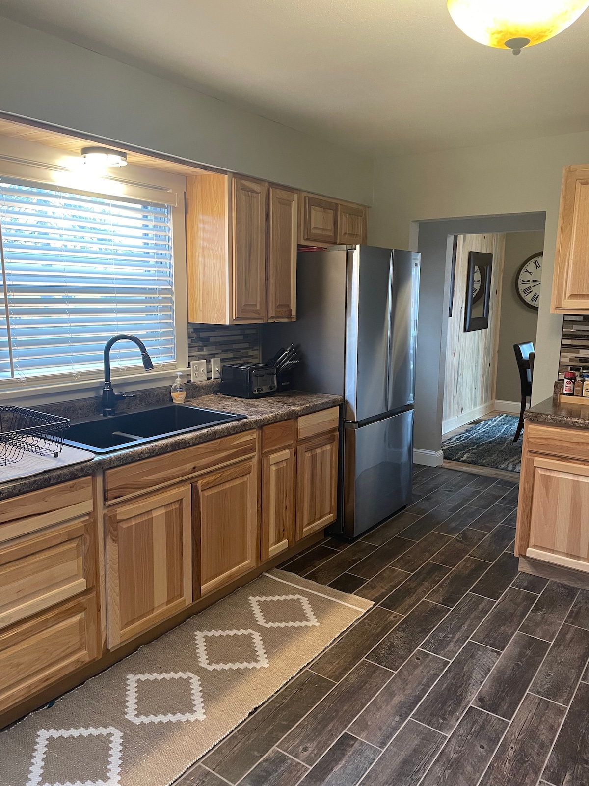 A modern kitchen is equipped with wooden cabinets and a stainless steel refrigerator. Counter space features a sink and decorative elements. Natural light enters through the window adorned with blinds. The dark tile floor adds a sleek contrast to the warm wood tones.
