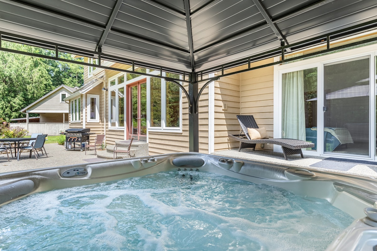 A hot tub is featured beneath a covered gazebo, providing a relaxing retreat surrounded by lush trees. The backdrop displays a spacious home with large glass doors, offering glimpses of the inviting outdoor seating area and additional accommodations.