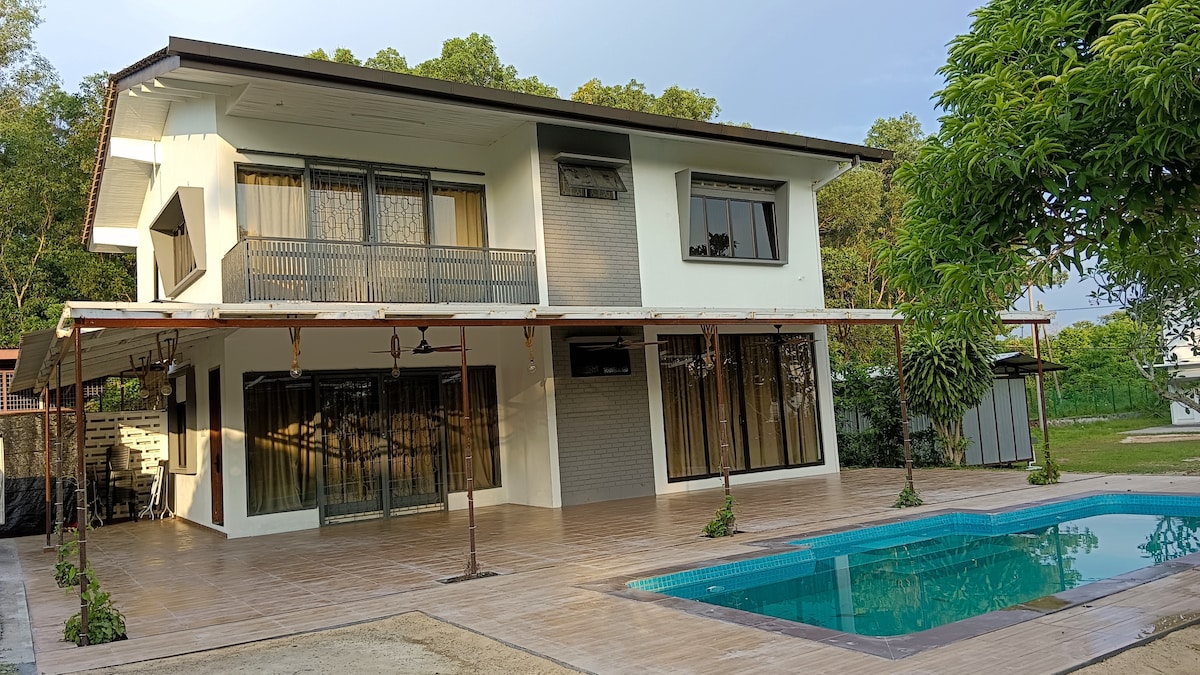 The two-story bungalow showcases a modern exterior with large glass windows and a spacious patio area. A turquoise swimming pool is set alongside the house, surrounded by landscaped greenery. Ample outdoor seating areas provide opportunities for relaxation.