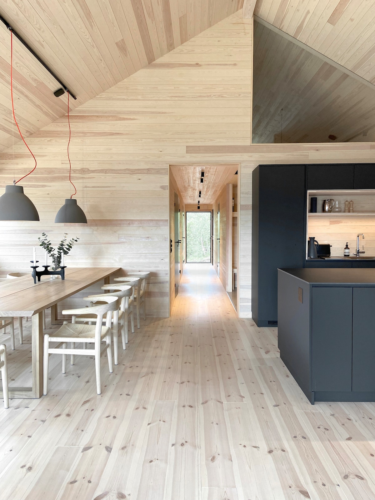 An open-concept interior features light wooden walls and flooring, showcasing a spacious dining area with a long table and white chairs. A modern kitchen with dark cabinetry is visible, along with pendant lighting and a narrow hallway leading to an outdoor view.
