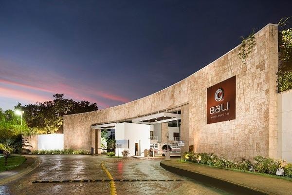 The entrance to a modern resort is presented, featuring an elegant stone facade with a prominent logo. The driveway is lined with greenery and illuminated softly by exterior lighting, providing a welcoming approach as the sun sets in the background.