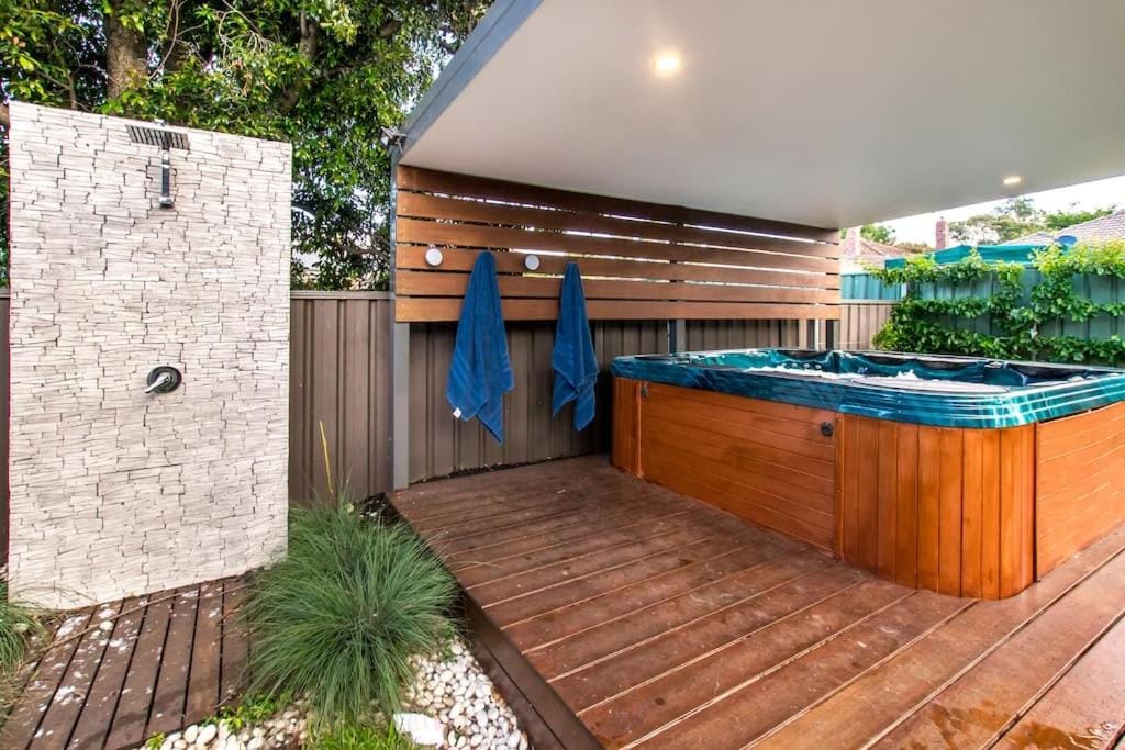An outdoor space features a wooden deck with a hot tub and a shower area. Two towels hang from the wall beside the shower, while greenery surrounds the space, adding a touch of nature. The design promotes relaxation and comfort.