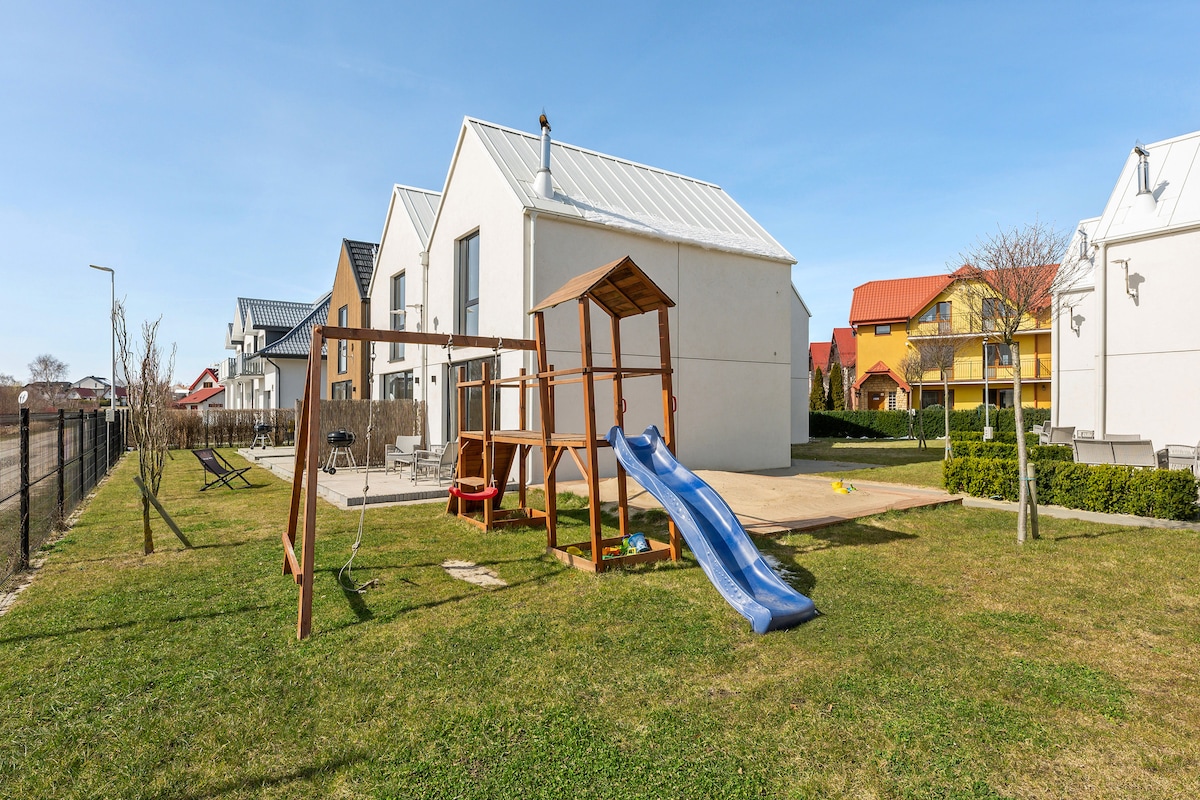 An outdoor space features a wooden playset with a slide and swing set, surrounded by a well-maintained grassy area. The backdrop includes several modern buildings with sloped roofs, under a clear blue sky.