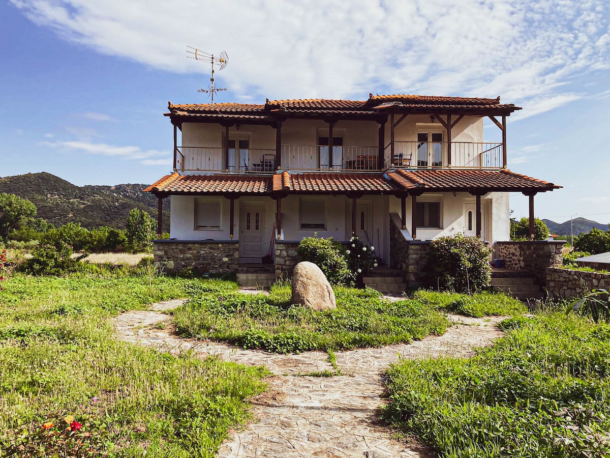 The country house features a two-story design with a tiled roof and wooden accents. A natural stone pathway leads to the entrance, surrounded by a lush green landscape. Balconies offer an outdoor view, while a large stone in the foreground adds a rustic element.