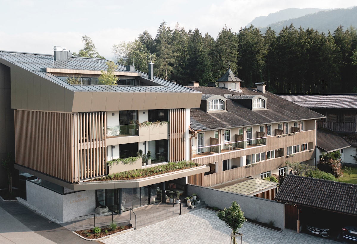A modern building features a sleek design with wooden and stone elements. Lush greenery surrounds the entrance, and large windows offer a view of the amenities. The structure is complemented by a backdrop of forested hills, creating a serene environment.
