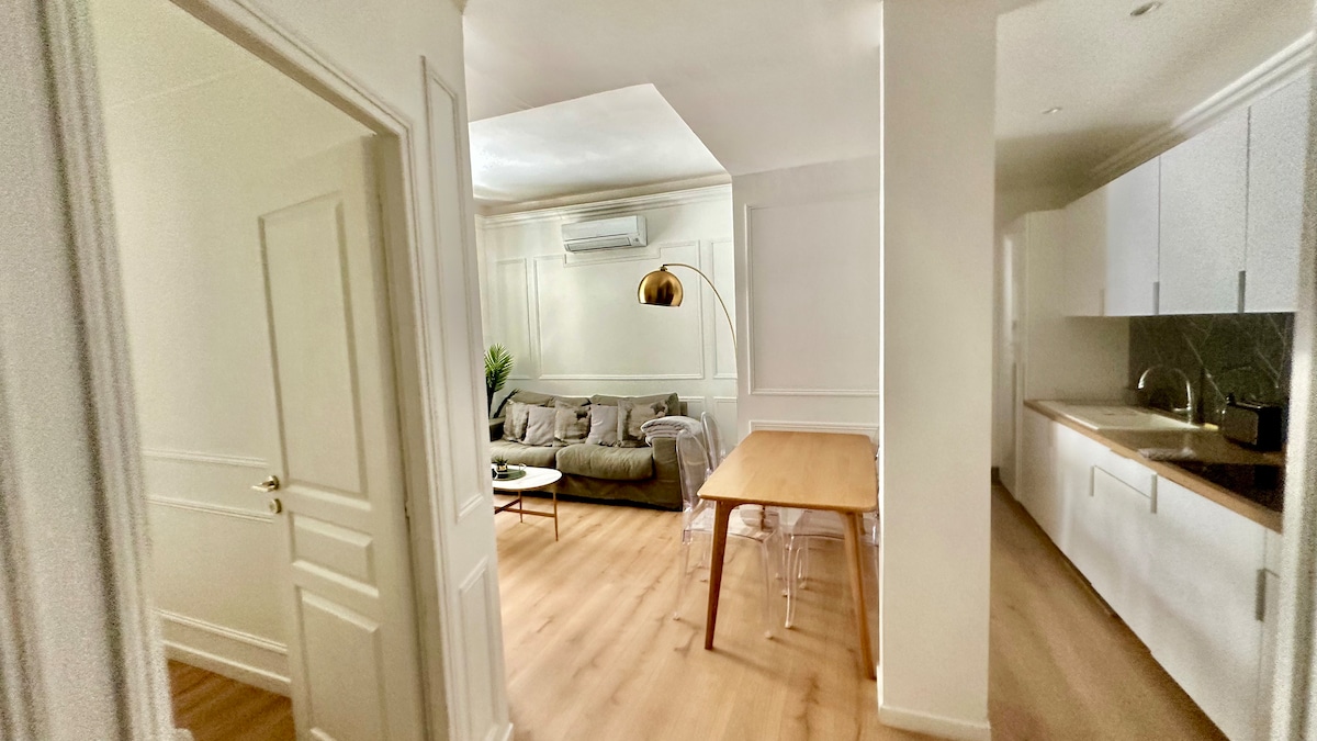 A light and airy hallway leads to an inviting living space. A comfortable gray sofa is paired with a stylish floor lamp, while a wooden dining table complements the open kitchen area, which features sleek cabinetry and modern appliances. Bright walls enhance the overall spaciousness.
