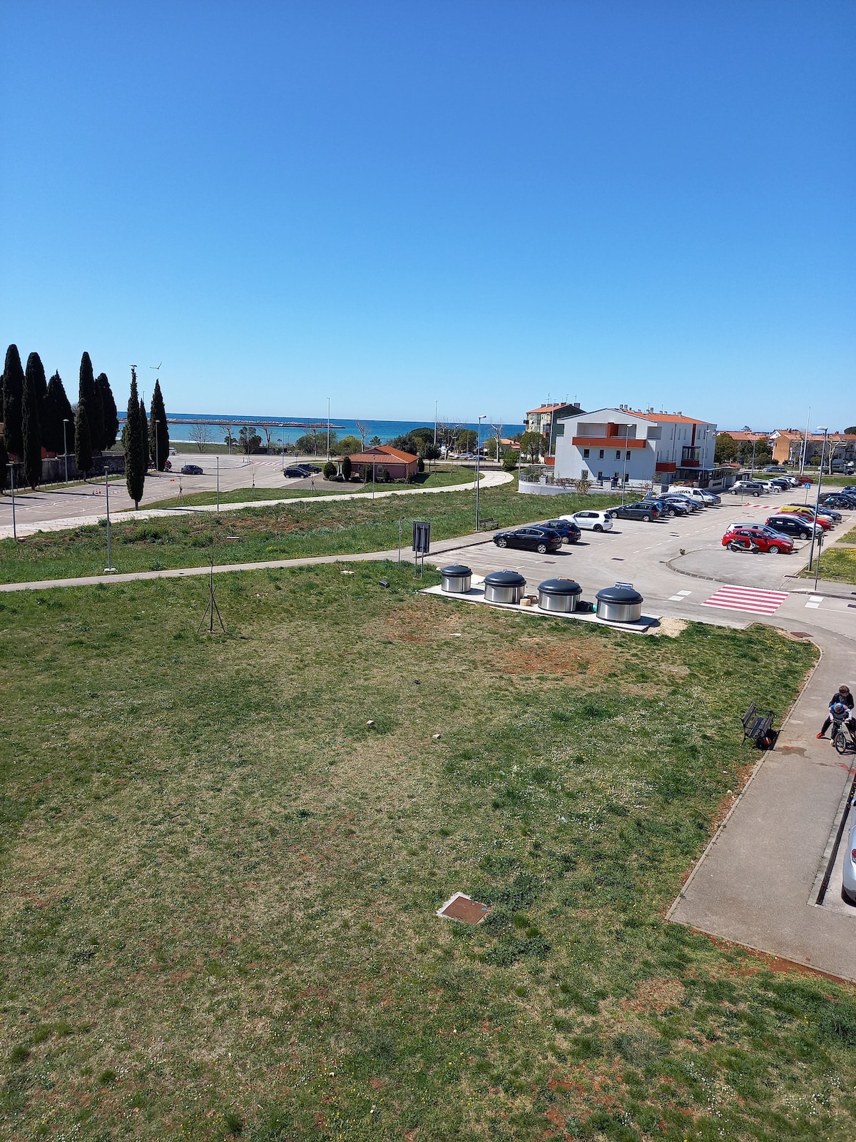 A view overlooking a grassy area and parking lot, with several parked cars visible. In the distance, the blue sea can be seen. The scene is complemented by tall trees lining the edge of the property, contributing to a serene outdoor setting.